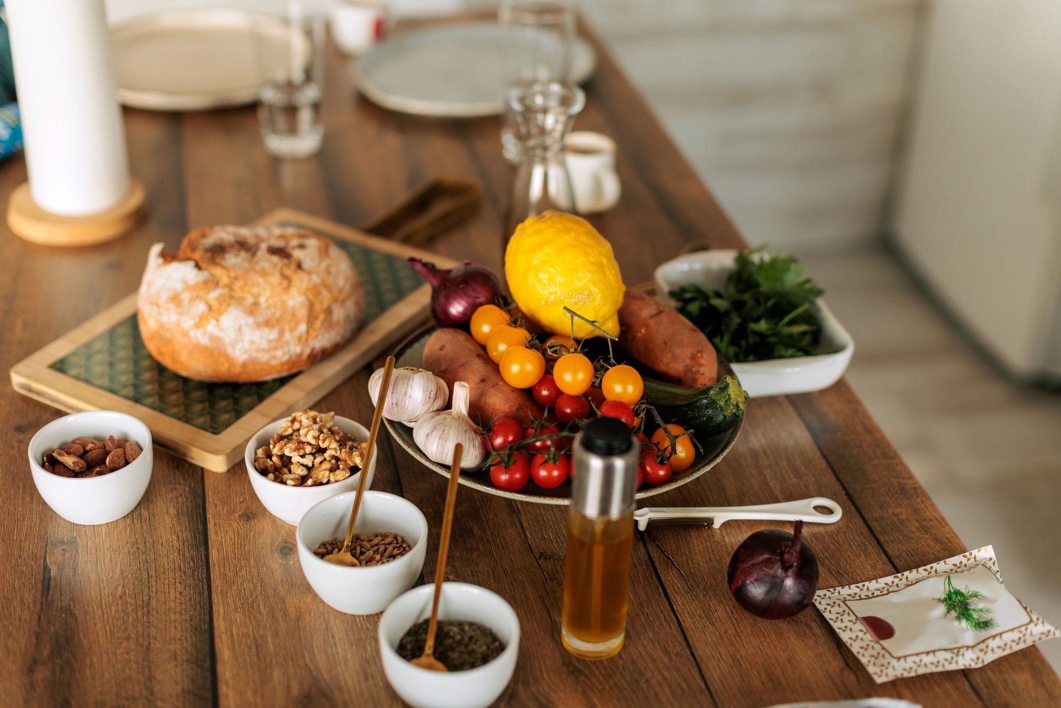 Rustic wooden table set with fresh garden vegetables, herbs, bread, nuts, and olive oil